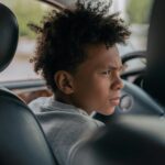 A teenager with curly hair sits inside a car, visibly concentrating as he imagines driving.