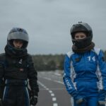 Two young drivers in racing suits and helmets standing on a go-kart track.